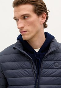 Marineblauwe gewatteerde jas met een hoge kraag en ritssluiting, gelaagd over een marineblauwe hoodie. Beschikt over een gestructureerde stof en een logo-accent op de borst.