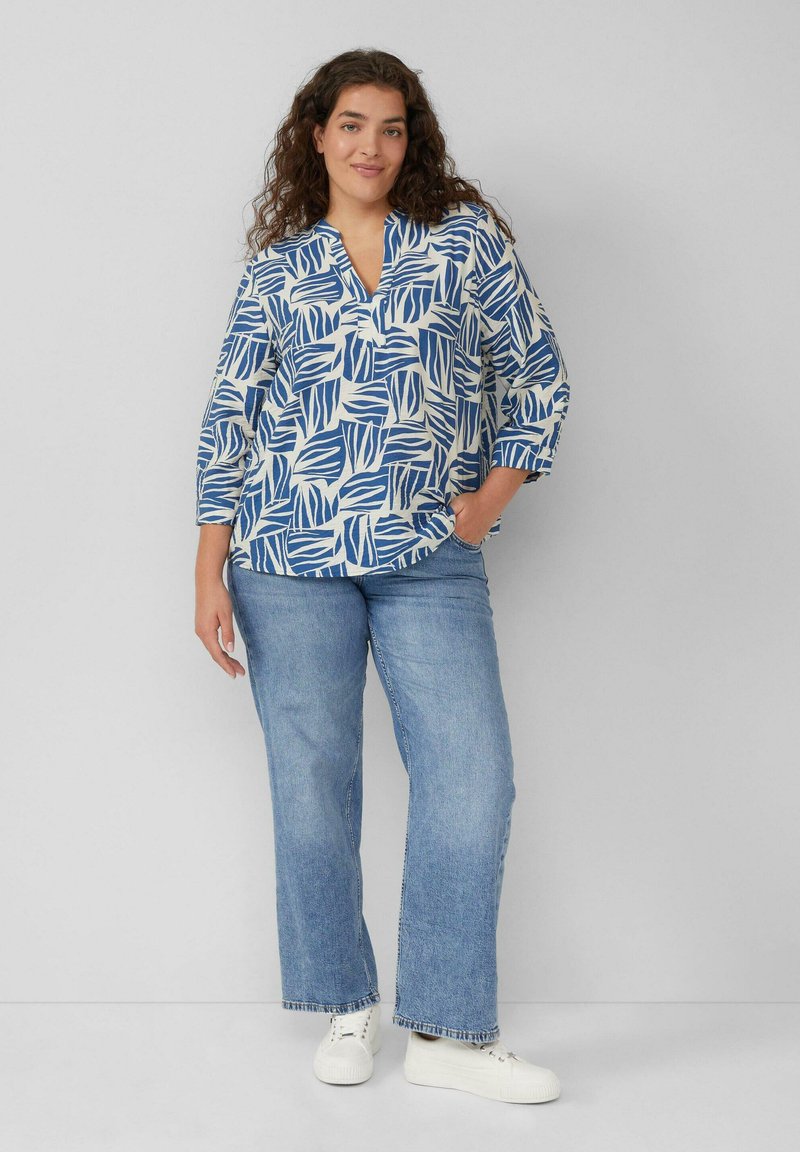 Witte top met blauw bladerenpatroon, V-halslijn, driekwart mouwen. Gepaarde met lichtblauwe wijde jeans en witte sneakers.