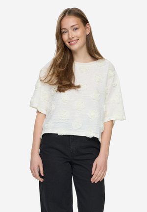 Młoda kobieta o długich brązowych włosach, ubrana w białą teksturowaną bluzkę z krótkim rękawem i czarne spodnie z wysokim stanem, uśmiechająca się do kamery.