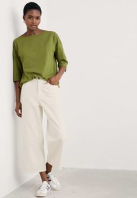 Olive green, loose-fit top with three-quarter sleeves paired with high-waisted, wide-leg cream trousers and white sneakers with purple accents.