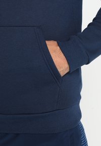 La mano di un uomo è appoggiata all'interno della tasca anteriore di una felpa blu navy con polsini e orlo a coste.