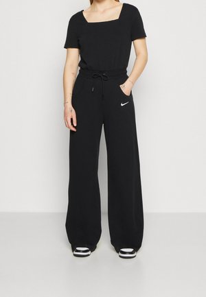 Vrouw draagt zwarte, wijd uitlopende Nike joggingbroek met aantrekkoord en een zwart kort mouwig topje met vierkante hals, staat met één hand in haar zak.