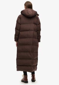 Lang, brun pufferjakke med hætte, der har et quiltet design, tekstureret materiale og sideslidser for bevægelighed. Satinlignende finish.