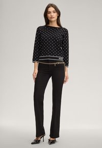 Μαύρο πουλόβερ με πουά σχέδιο και μανίκια τριών τετάρτων, συνδυασμένο με μαύρο παντελόνι και ζώνη αλυσίδα, που διαθέτει λογότυπο στη μέση.