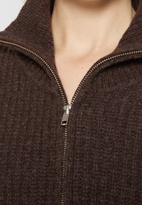 Gros plan sur un pull marron tricoté avec une fermeture éclair argentée partiellement ouverte et un col visible sur une peau claire.