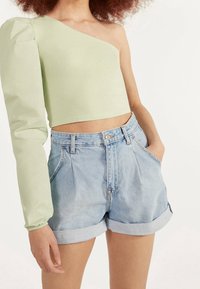 Vaaleanvihreä croppitoppi, jossa on yksi pitkä puffihihna, yhdistetty korkeavyötäröisiin farkkushortseihin, joissa on käännetyt hihansuut ja haalistunut sininen väri.