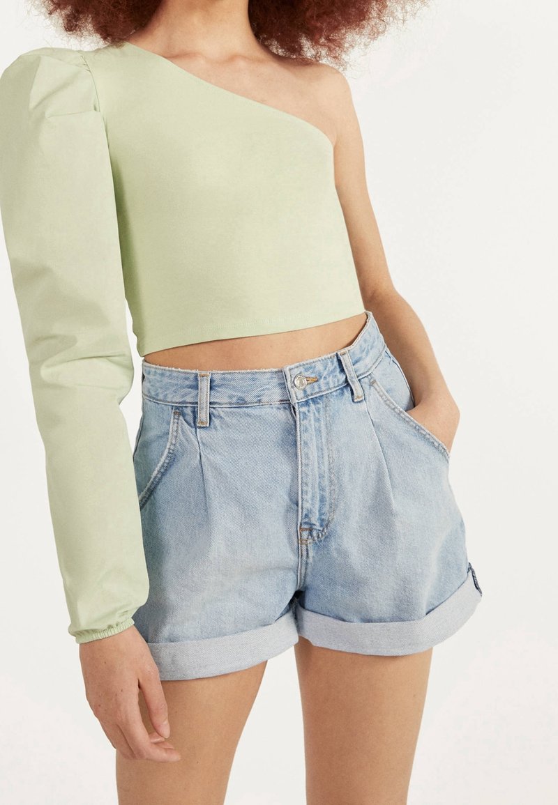 Vaaleanvihreä croppitoppi, jossa on yksi pitkä puffihihna, yhdistetty korkeavyötäröisiin farkkushortseihin, joissa on käännetyt hihansuut ja haalistunut sininen väri.