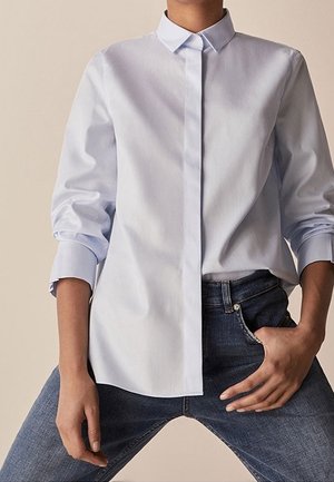 Person iført en lyseblå, langærmet, knaplukket skjorte med op-rullede ærmer og mørkeblå jeans, poserende med den ene hånd i lommen.