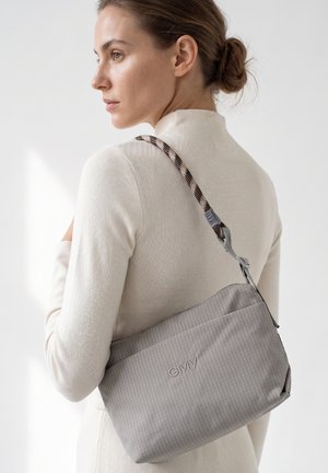Donna con i capelli raccolti in uno chignon che indossa un dolcevita color crema porta una borsa a tracolla grigia a righe sottili con una tracolla decorata sulla schiena.