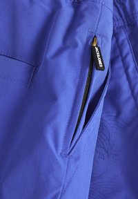 Shorts en tissu violet avec une poche zippée, une taille élastique et un motif subtil imprimé sur un côté. Étiquette de la marque visible près de la fermeture éclair.