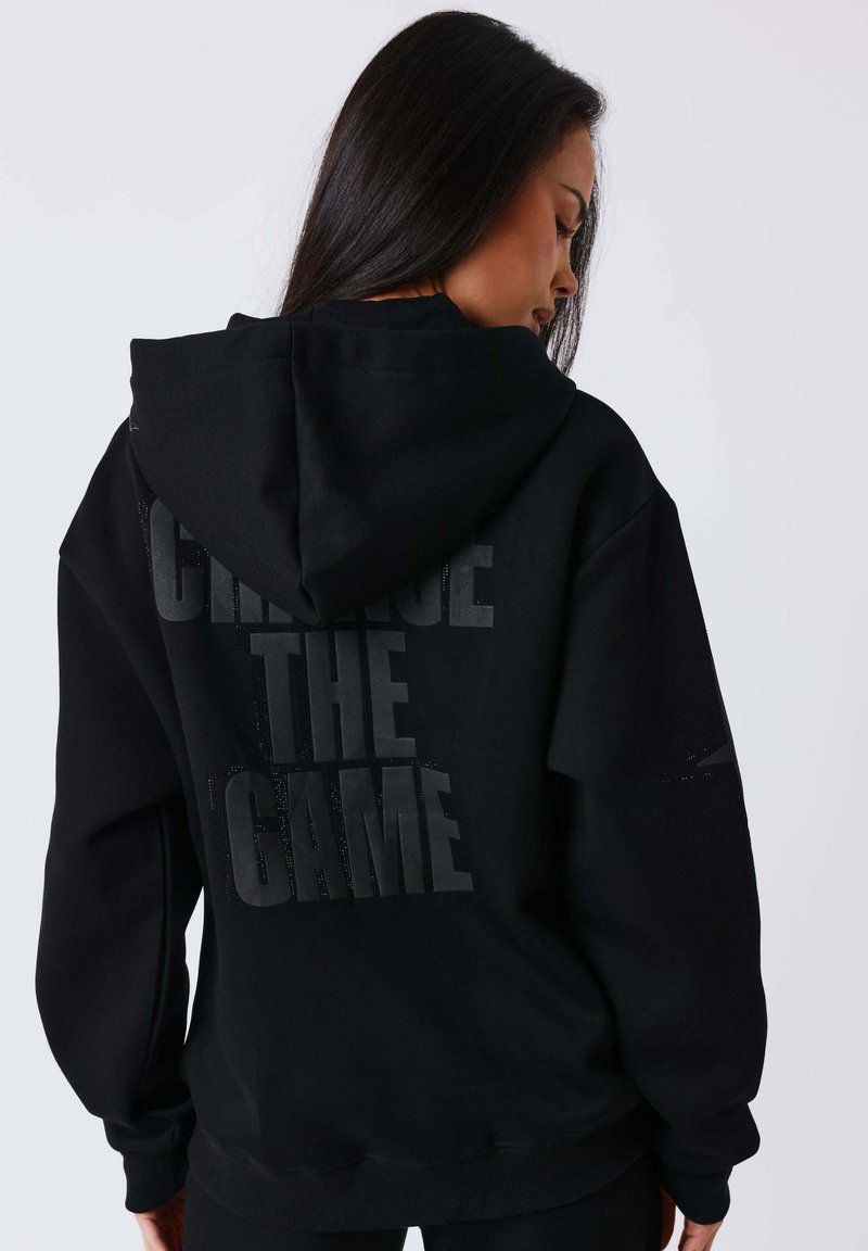 Mujer con cabello largo y oscuro de perfil, vistiendo una sudadera negra con capucha puesta y la frase "CAMBIA EL JUEGO" impresa en la espalda con letras oscuras.