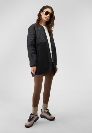 Frau trägt eine übergroße schwarze gesteppte Jacke, braune Leggings, beige Socken, pinke Wanderschuhe und große verspiegelte Sonnenbrille, steht vor grauem Hintergrund.
