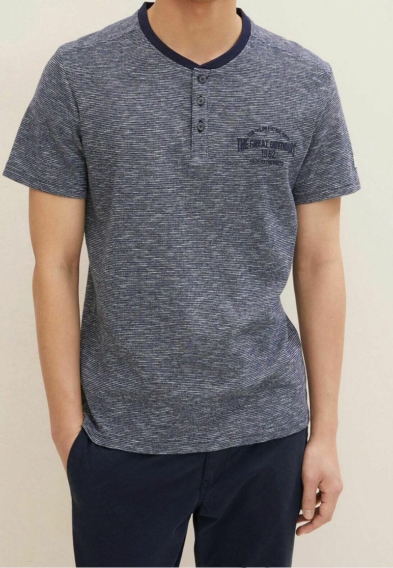 Námořnická modrá pánská tričko s krátkým rukávem a decentním proužkovaným vzorem, kulatým výstřihem, dvěma knoflíky a tištěným textem na hrudi.