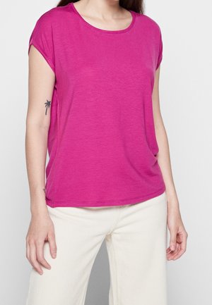 Vrouw die een magenta shirt met korte mouwen en witte broek draagt, met een kleine palmboomtatoeage zichtbaar op haar linkerarm.