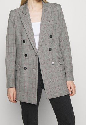 Kvinde iført en grå ternet dobbeltradet blazer med sorte knapper, hvid top og sorte bukser, stående mod en ensfarvet baggrund.