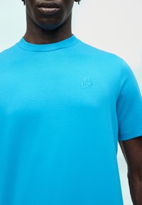 Άνδρας που φοράει φωτεινό μπλε μπλουζάκι με στρογγυλή λαιμόκοψη και διακριτικό κεντητό προφίλ λογότυπο στο αριστερό στήθος, με ανοιχτό φόντο.