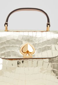 Złota torebka z wytłoczeniem krokodyla, z górnym uchwytem, z świecącym złotym logo w kształcie łopaty i gładkimi skórzanymi akcentami.