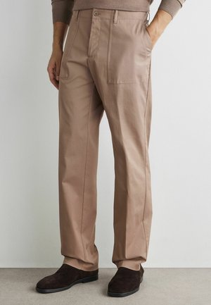 Homme portant un pantalon beige droit avec des poches plaquées à l’avant et des chaussures en daim marron foncé, debout avec une main dans la poche.
