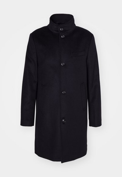 Tamsiai mėlynas vilnonis paltas su aukštu apykakle, vieno mygtuko dizainu ir penkiomis didelėmis mygtukais. Turi dvi šonines kišenes ir krūtinės kišenę.