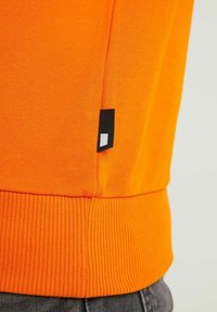 Primo piano dell'orlo di un maglione a coste di un vivace arancione, con un piccolo etichetta nera che presenta un design rettangolare bianco, indossato sopra pantaloni grigio scuro.