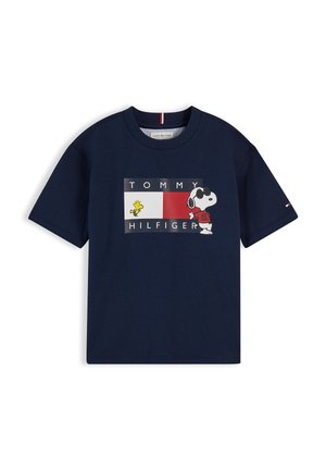 Jeune fille avec deux chignons portant un t-shirt Tommy Hilfiger bleu marine avec Snoopy, un pantalon large beige et des baskets blanches, posant sur fond blanc.