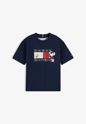 Jeune fille avec deux chignons portant un t-shirt Tommy Hilfiger bleu marine avec Snoopy, un pantalon large beige et des baskets blanches, posant sur fond blanc.