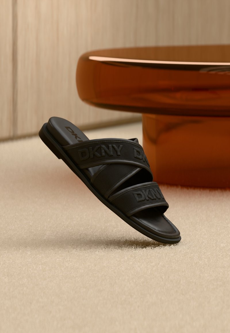 Mule DKNY noire avec deux brides et logo embossé, suspendue en l'air au-dessus d'un tapis beige près d'une table ronde ambrée.