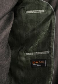 Innenseite einer Jacke mit einem strukturierten dunkelgrauen Stoff, einem grün gemusterten Futter, einer kleinen Tasche und einem Etikett mit der Aufschrift "3D WOLLE."