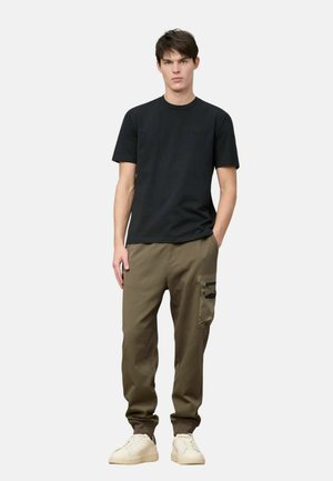 Giovane uomo in piedi con le mani in tasca, indossa una camicia nera a maniche corte, pantaloni cargo color oliva e sneakers bianche su sfondo bianco.