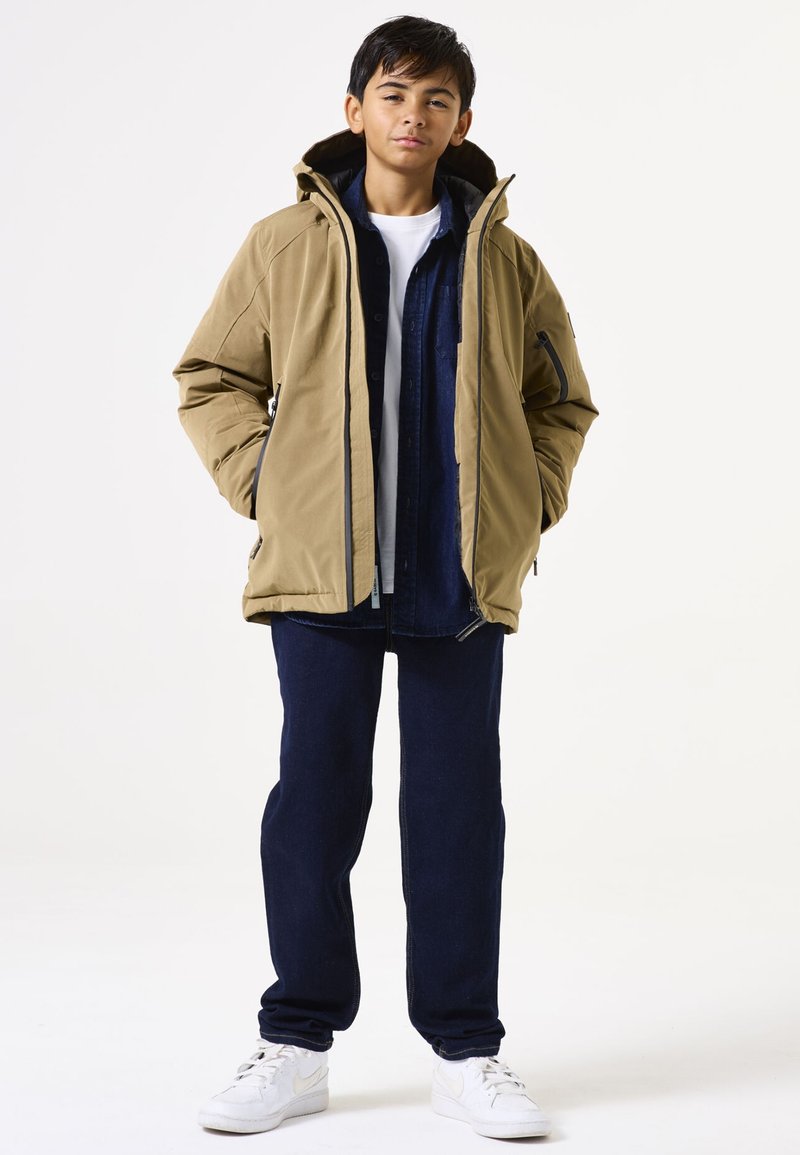 Veste isolante beige avec capuche, portée sur une chemise à boutons sombre et un t-shirt blanc. Associée à un pantalon bleu marine et des baskets blanches.