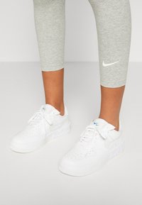 Zapatillas Nike Air Force 1 blancas con puntera perforada, cordones planos y acento en azul claro en el swoosh, combinadas con leggins gris hasta la pantorrilla.