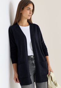 Cardigan navy in materiale morbido, con apertura frontale, maniche lunghe e due tasche frontali. Abbinato a un top bianco e jeans grigi.