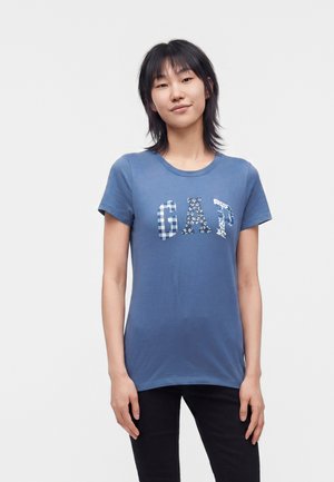 Jeune femme aux cheveux courts noirs portant un t-shirt bleu avec des lettres à motifs formant le mot GAP et un pantalon noir, debout devant un fond uni.