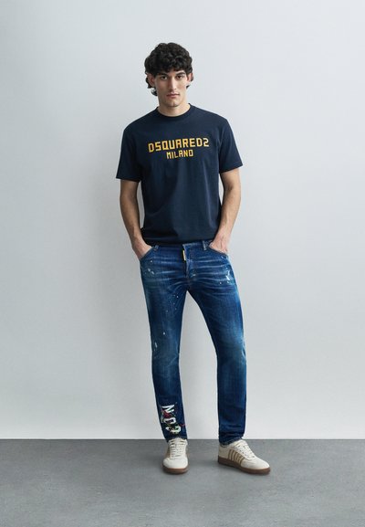 Marineblå t-shirt med gul tekst, kombineret med slidte blå jeans med farverige lapper. Hvide sneakers med lysebrune såler.