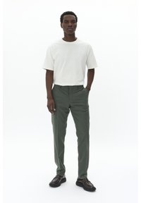 T-shirt blanche à manches courtes, pantalon vert ajusté et chaussures marron foncé. L'ensemble est décontracté et les matériaux sont lisses, sans motifs visibles.