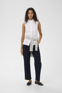 Hvid ærmeløs skjorte med høj krave, kombineret med mørkeblå bukser med vidde og en lysegrå trøje bundet om livet. Sorte sko.