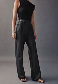 Zwarte, hooggesneden nepleren broek met brede pijpen, voorzien van voornaad en een gladde textuur. Gecombineerd met een aansluitende zwarte top.