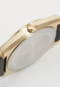 Gold-tone watch with a rounded case, featuring a textured crown marked "HUGO," and a smooth black strap. Water-resistant design.