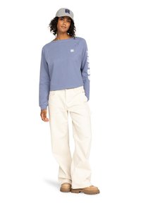 Blaues, cropped Sweatshirt mit weißem Blumenmuster an den Ärmeln, kombiniert mit cremefarbenen, weiten Hosen und braunen Plateauschuhen. Graue Kappe mit dem Buchstaben R.