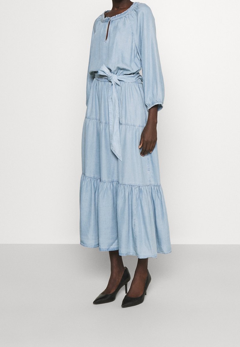 Ljusblå denimklänning med lång ärm, volang vid halsen och flerlagad kjol. Inkluderar ett matchande bälte i midjan och bärs med svarta klackskor.