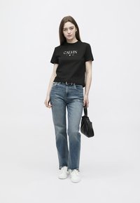 Femeie purtând un tricou negru Calvin, blugi albaștri, adidași albi, ținând o geantă mică neagră, stând pe un fundal alb simplu.