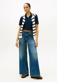 Granatowa koszulka polo zestawiona z prążkowanym bezrękawnikiem, szerokimi, jasnoniebieskimi dżinsami i brązowymi loaferami. Świeże, fakturowane tkaniny.