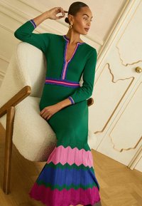 Rochie tricotată verde cu mâneci lungi, decolteu cu margine ondulată, model chevron roz și albastru la tiv și garnitură contrastantă la mâneci și talie.