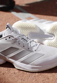 Witte sportschoenen met lichtgrijze strepen en gestructureerde zolen die rusten op een roodachtige ondergrond.