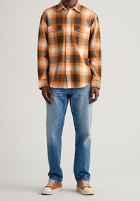 Camicia flanella a quadri arancioni e marroni con due tasche sul petto, abbinata a jeans chiari strappati e scarpe da ginnastica marroni.