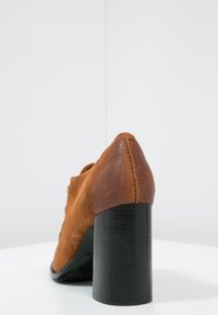 Zapato de tacón alto de ante marrón con un acabado texturizado y un robusto tacón negro. La forma es alargada con una punta puntiaguda.