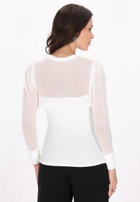 Maglietta bianca a maniche lunghe con schiena trasparente, adornata con piccole perline, dettagli in tessuto arricciato e design aderente. Texture liscia e vestibilità slim.