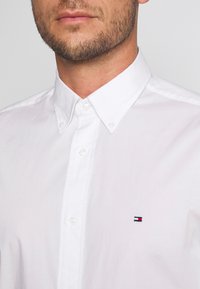Uomo che indossa una camicia bianca a maniche lunghe con un piccolo logo rosso, bianco e navy sul petto e una barba ben curata.