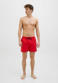 Bañador rojo hecho de material ligero, con un cordón negro en la cintura y texto blanco a lo largo del costado. Diseño simple, sin patrones.