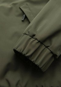 Primo piano di una manica di giacca verde oliva con polsino elastico e una piccola cerniera nera su una tasca sullo sfondo.